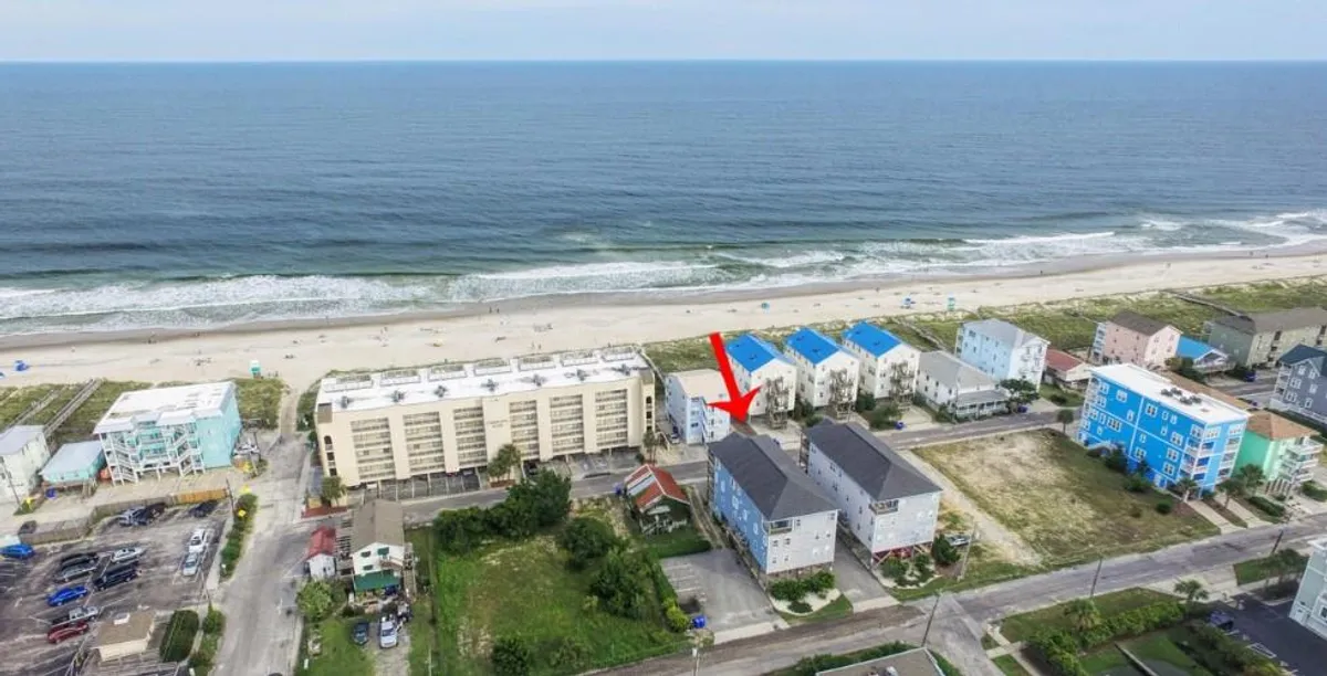 A tranquil exterior view showing the ocean, sandy beach, neighboring buildings, and surrounding green spaces. The wide setting includes hints of activity spaces suitable for family gatherings and leisure walks.