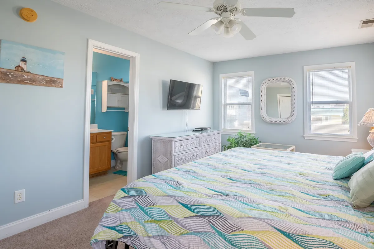 A bright and spacious bedroom featuring a king bed adorned with a colorful quilt, a wall-mounted TV, a decorative mirror, and large windows letting in ample natural light. The space feels comforting and serene, perfect for relaxation after a long day.