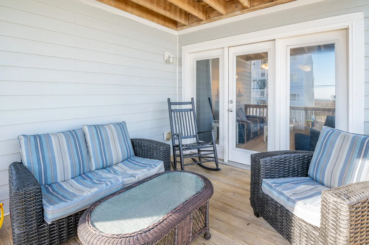 This image captures a comfortable exterior deck space with inviting wicker seating, striped cushions, a glass-top coffee table, and sliding glass doors opening to the interior. The arrangement suggests a place for relaxing with a book, having morning coffee, or enjoying time with friends. The setup directly matches the described features in the section, such as outdoor sofa, coffee table, and an atmosphere of comfort and tranquility.