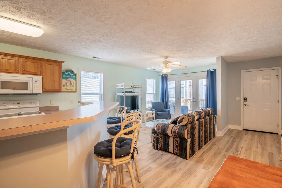 This image captures an open-concept living area where a comfortable sofa, kitchen counter with bar stools, blue curtains, and a dining table are visible. The layout clearly shows the seamless flow between the living, dining, and kitchen spaces. Natural light streams in, enhancing the casual, welcoming ambiance.