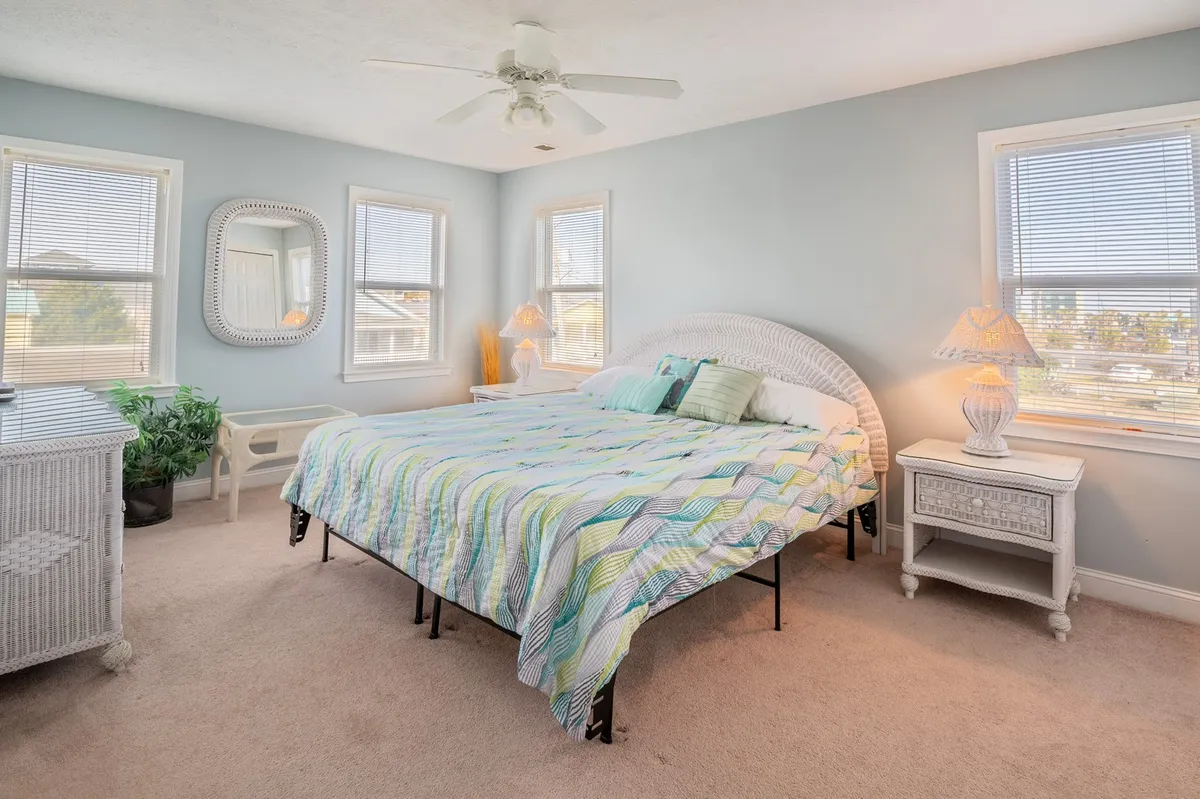A bedroom filled with natural light, featuring a colorful quilt, white wicker furniture, and large windows. The atmosphere is peaceful and refreshing, ideal for quiet mornings with a view.