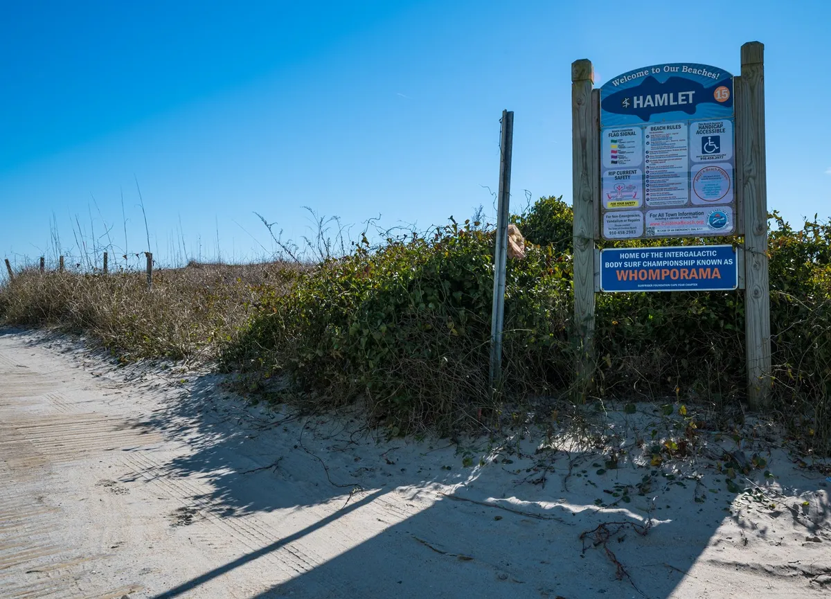 A sandy path leading from coastal vegetation to the beach, marked by a welcoming beach sign, suggesting easy access and the start of a day of seaside adventures.