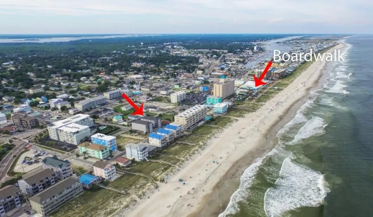 A sweeping view of the Carolina Beach Boardwalk area with beachfront properties in the foreground, the iconic boardwalk extending along the sand, the ocean's surf, and families walking or visiting local shops and restaurants.