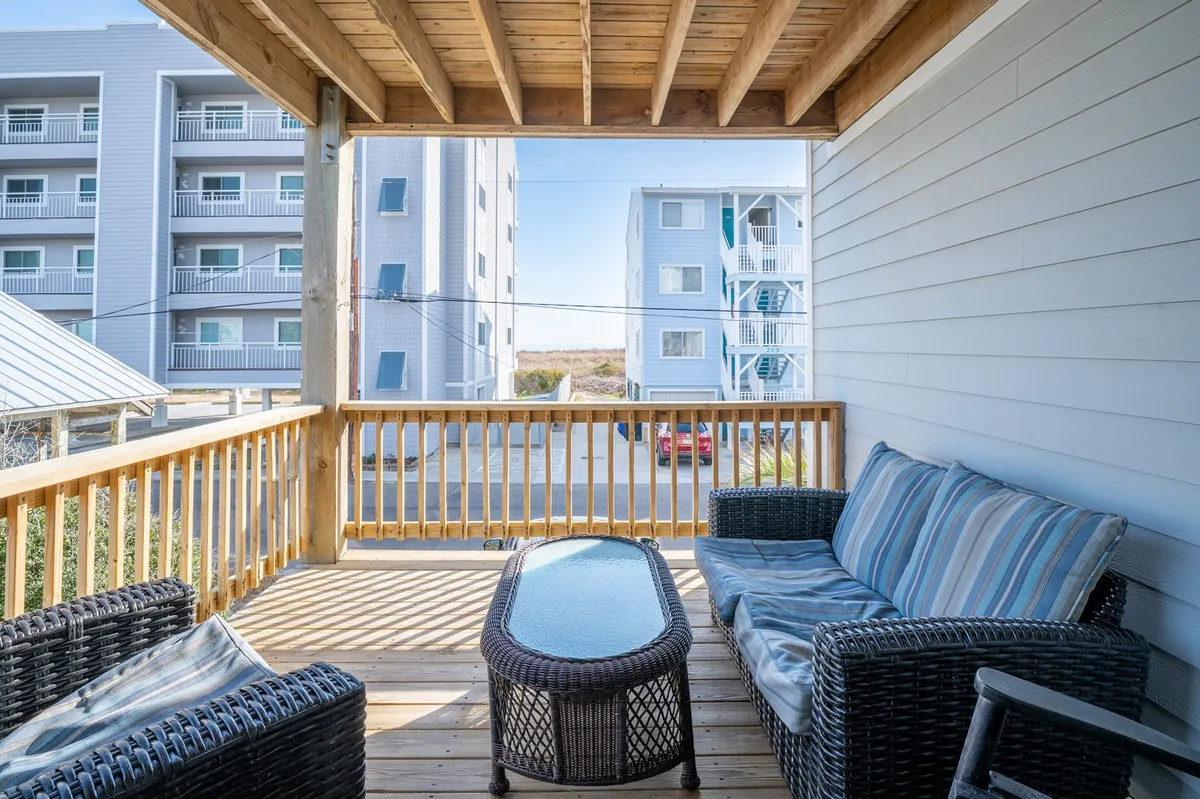 An exterior shot of an outdoor seating area on a deck, with a view through the railing to surrounding buildings. The scene conveys relaxation and sociability, suggesting the deck as a space for morning coffee, reading, or gatherings with friends.