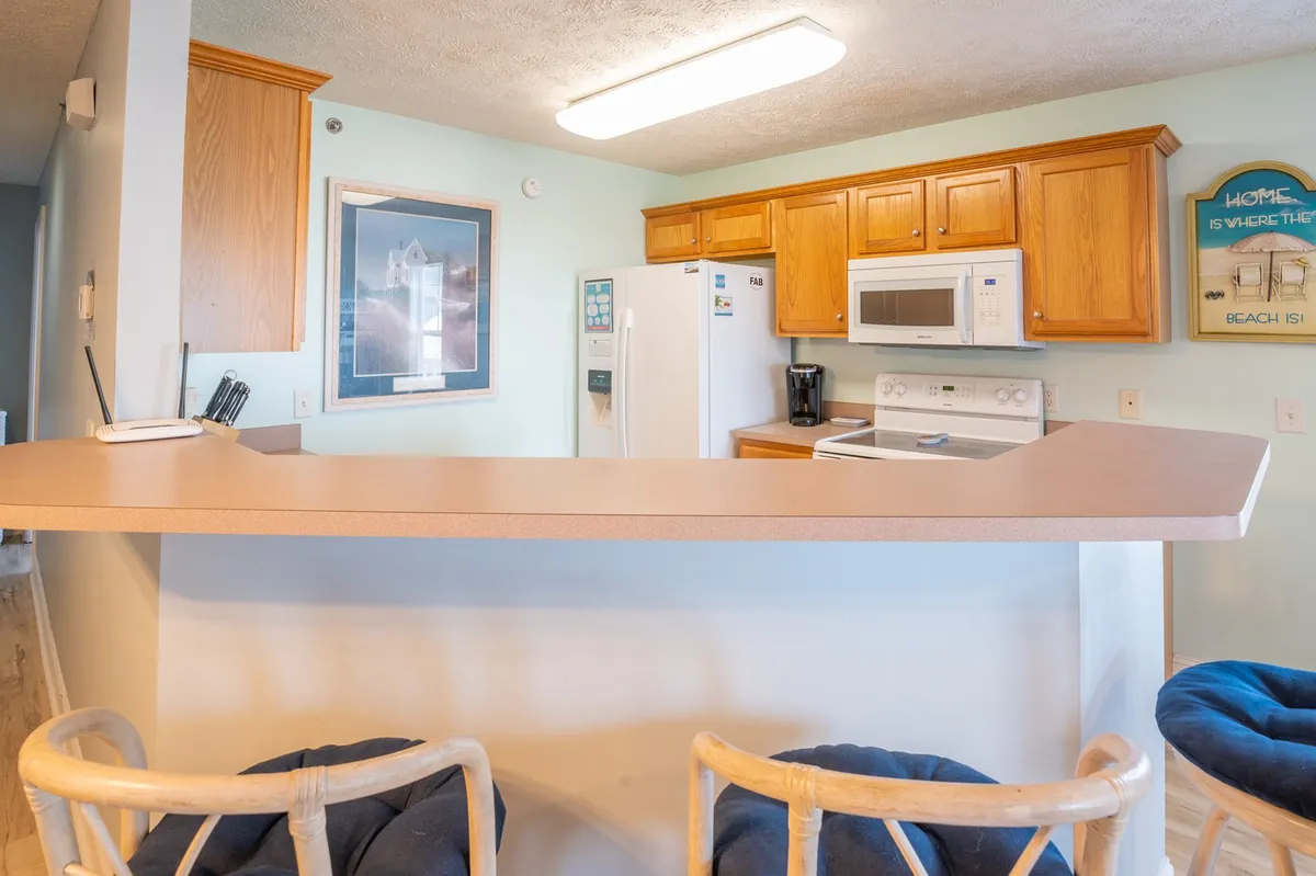 A view of the inviting kitchen with a prominent island, wooden cabinets, white appliances, framed artwork, and bar stools. The space invites socializing and casual meals, with a clear view of the breakfast bar and opportunities for meal prep and conversation.