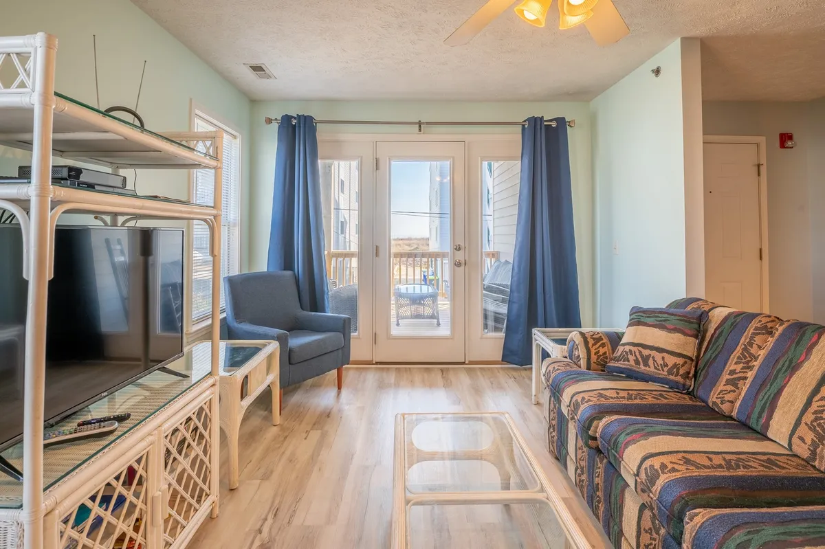 A bright living area featuring a cozy sofa facing a television, with sliding glass doors that open to outside views. The space is bathed in natural light, evoking a breezy, comfortable feel ideal for relaxing or movie nights.