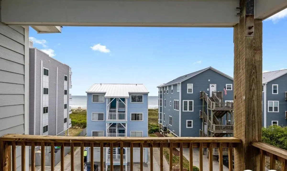 This image captures a peaceful balcony scene with blue buildings in the distance, a prominent balcony railing, and sweeping ocean views. The scene suggests a relaxing atmosphere where guests can unwind, sip coffee, and enjoy ocean breezes or sunsets.