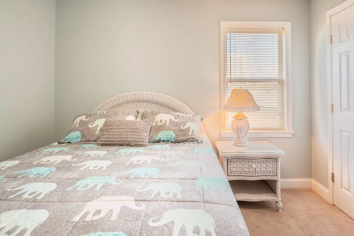 A welcoming queen bedroom featuring elephant-patterned bedding, a softly glowing lamp, and decorative pillows. The space is serene and inviting, ideal for relaxation or curling up with a book.