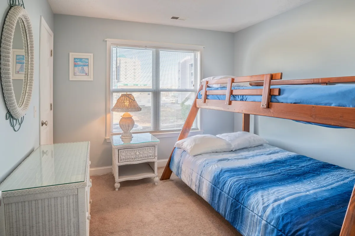A playful bunk room with a twin over full bunk bed, a window for natural light, a nightstand with a lamp, and a decorative mirror. The mood is relaxed and comfortable, making it ideal for kids or teens to unwind, read, or play.